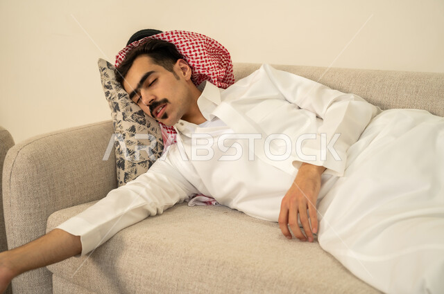 الاحساس بالكسل والخمول والحاجة إلى الاسترخاء، النوم بعمق من الارهاق والتعب، رجل عربي خليجي سعودي يرتدي الثوب والشماغ يقوم بالاستلقاء على الاريكة في منزله، اخذ قيلولة و قسط من الراحة، الاستمتاع بالأجواء الهادئة