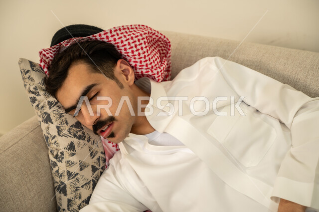 اخذ قيلولة و قسط من الراحة، الاحساس بالكسل والخمول والحاجة إلى الاسترخاء، النوم بعمق من الارهاق والتعب، صورة مقربة لرجل عربي خليجي سعودي يرتدي الثوب والشماغ يقوم بالاستلقاء على الاريكة في منزله، الاستمتاع بالأجواء الهادئة