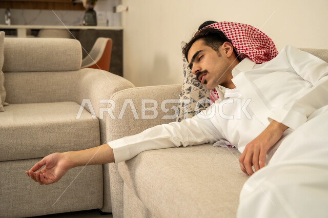النوم بعمق من الارهاق والتعب، رجل عربي خليجي سعودي يرتدي الثوب والشماغ يقوم بالاستلقاء على الاريكة في منزله، اخذ قيلولة و قسط من الراحة، الاحساس بالكسل والخمول والحاجة إلى النوم، الاسترخاء والاستمتاع بالأجواء الهادئة