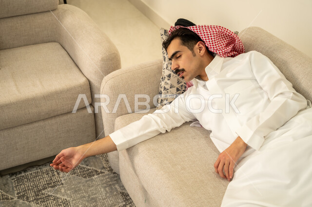 الاحساس بالكسل والخمول والحاجة إلى الاسترخاء، النوم بعمق من الارهاق والتعب، رجل عربي خليجي سعودي يرتدي الثوب والشماغ يقوم بالاستلقاء على الاريكة في منزله، اخذ قيلولة و قسط من الراحة، الاستمتاع بالأجواء الهادئة
