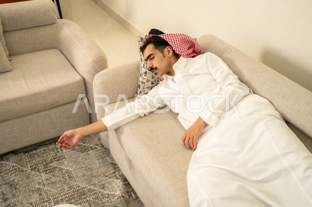 النوم بعمق من الارهاق والتعب، رجل عربي خليجي سعودي يرتدي الثوب والشماغ يقوم بالاستلقاء على الاريكة في منزله، اخذ قيلولة و قسط من الراحة، الاحساس بالكسل والخمول والحاجة إلى النوم، الاسترخاء والاستمتاع بالأجواء الهادئة