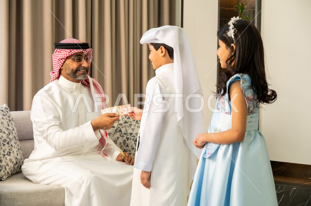 الاحتفال مع الابناء في ايام العيد السعيد في السعوديه، إيماءات الفرح والسرور بالأحفاد، صلة الرحم والترابط الاسري, حسن اكرام واستقبال الضيف, التجمعات والزيارات الاسرية في الأعياد والمناسبات الاسلامية المباركة, رجل عربي خليجي سعودي يقوم بتقديم العيدية لحفيده