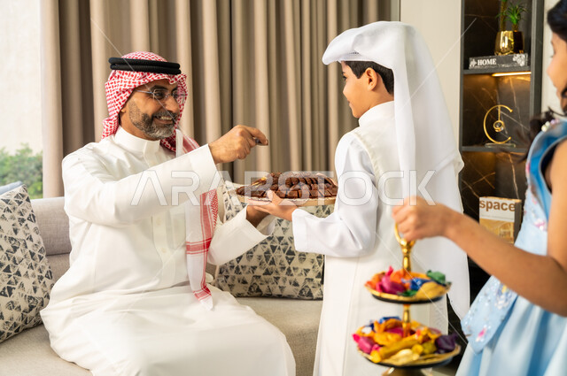 التجمعات والزيارات الاسرية في الأعياد والمناسبات الاسلامية المباركة, ولد عربي خليجي سعودي يرتدي الزي التقليدي يقوم بتقديم الحلوى لجده، أجواء العيد في المجتمع العربي، إيماءات الفرح والسرور، مفهوم صلة الرحم والترابط الاسري, حسن اكرام واستقبال الضيف