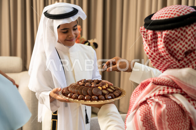 أجواء العيد في المجتمع العربي، إيماءات الفرح والسرور، مفهوم صلة الرحم والترابط الاسري, حسن اكرام واستقبال الضيف, التجمعات والزيارات الاسرية في الأعياد والمناسبات الاسلامية المباركة, ولد عربي خليجي سعودي مبتسم يرتدي الزي التقليدي يقوم بتقديم الحلوى لجده