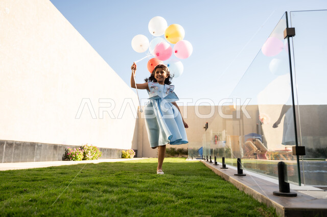 Showing feelings of joy and happiness at the arrival of Eid, celebrating holidays and occasions outdoors, a Saudi Arabian Gulf girl wearing a dress holding a group of colorful balloons in her hand running in the garden of the house, the Eid atmosphere in Saudi Arabian society
