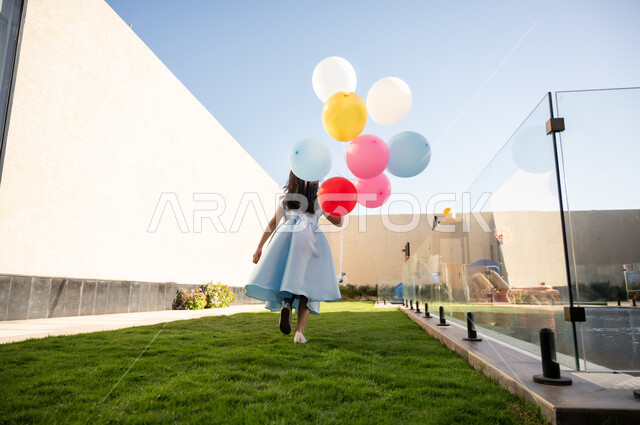 أجواء العيد في المجتمع العربي السعودي، الاحتفال في الأعياد والمناسبات في الهواء الطلق, إظهار مشاعر السرور والفرح بقدوم العيد, صورة من الخلف لفتاة عربية خليجية سعودية ترتدي فستان تمسك بيدها مجموعة من البلالين الملونة تركض في حديقة المنزل