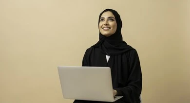 مهنة ووظيفة نسائية مكتبية سعودية، ادارة وانجاز المهام باستخدام جهاز تقني حديث ومتطور, بورتريه مقرب لإمرأة عربية خليجية سعودية مبتسمة ترتدي الحجاب والعباءة تعمل على الحاسوب المحمول، ايماءات وتعابير السرور والسعادة، خلفية بيج