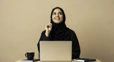  مهنة ووظيفة نسائية مكتبية سعودية، ادارة وانجاز المهام باستخدام جهاز تقني حديث ومتطور, بورتريه مقرب لإمرأة عربية خليجية سعودية مبتسمة ترتدي الحجاب والعباءة تعمل على الحاسوب المحمول، ايماءات وتعابير السرور والسعادة، خلفية بيج