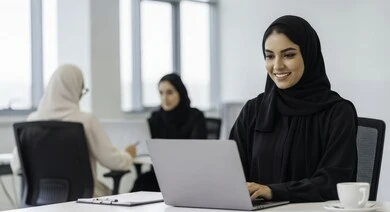 مهنة ووظيفة نسائية مكتبية سعودية، ادارة وانجاز المهام باستخدام جهاز تقني حديث ومتطور, إمرأة عربية خليجية سعودية مبتسمة ترتدي الحجاب والعباءة تعمل على الحاسوب المحمول في مقر الشركة، ايماءات وتعابير السرور والسعادة