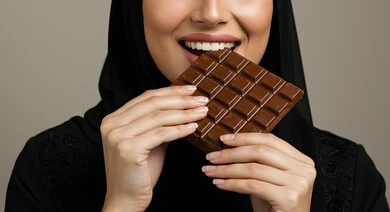 قطع الحلوى اللذيذة، سكريات و أطعمه ذات سعرات حراريه عاليه، بورتريه مقرب لإمرأة عربية خليجية سعودية ترتدي الحجاب والعباءة تقم بتناول لوح من الشوكولاته، قالب الكاكاو الشهية, خلفية رمادية
