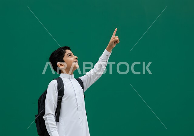 الاشارة الى الاعلى بتعابير السرور، بدء العام الدراسي  في المدارس السعودية، بورتريه من الجانب لتلميذ عربي خليجي سعودي يرتدي الثوب التقليدي يحمل حقيبة الظهر المدرسية يشير الى شي ما، العودة الى الفصل الدراسي بايماءات الحماس والسرور، خلفية خضراء
