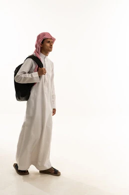 The concept of back to school, gestures of walking and going to a specific place, the concept of striving to achieve success, educational scientific curricula, a portrait of an Arab Gulf Saudi student wearing a white thobe and a shemagh carrying a backpack looking with gestures of happiness and joy, a full-body image, white background.