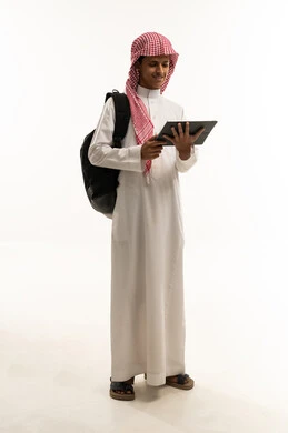 اللعب على الجهاز اللوحي،  قضاء وقت ممتع على الإنترنت، استخدام الأجهزة التقنية الحديثة، مفهوم التعليم وإنجاز المهام عن بعد، بورتريه لطالب عربي سعودي خليجي يرتدي الثوب التقليدي و الشماغ و حقيبة الظهر يقوم باستخدام التابلت، الجسم بالطول الكامل، خلفية بيضاء
