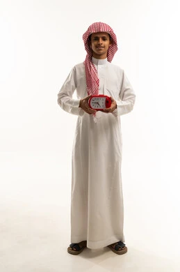 أهمية الاستيقاظ باكرا للذهاب إلى المدرسة، بورتريه لولد عربي خليجي سعودي يرتديالثوب الابيض و الشماغ يمسك ساعة المنبه بيده و ينظر الى الكاميرا بايماءات السعادة و السرور ، موسم العودة للمدارس في المملكة العربية، الجسم بالطول الكامل، خلفية بيضاء