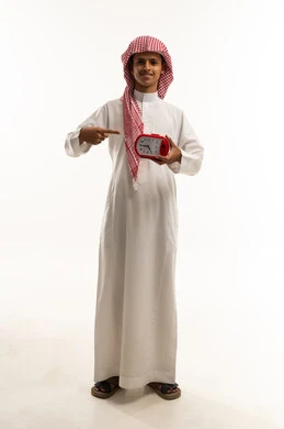 مفهوم تنظيم و إدارة الوقت, الاستعداد للعام الدراسي الجديد, بورتريه لولد عربي خليجي سعودب يرتدي الثوب الابيض و الشماغ يمسك ساعة المنبه بيده و يشير اليها, ايماءات السعادة و السرور، الجسم بالطول الكامل, خلفية بيضاء