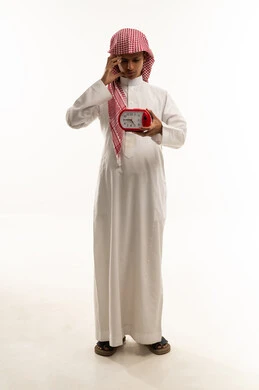 The need for rest and sleep, the concept of back to school, education in the Kingdom of Saudi Arabia, a portrait of an Arab Gulf Saudi boy wearing a white thobe and a shemagh holding an alarm clock in his hand, placing his hand over his eye with sleepy expressions, full body, white background.