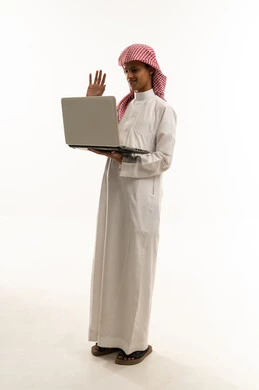 تعابير الترحيب والاستقبال، إجراء مكالمة فيديو باستخدام الحاسب المحمول، مفهوم التعليم عن بعد، بورتريه لصبي عربي خليجي سعودي يرتدي الثوب التقليدي و الشماغ يستخدم الحاسب المحمول في البحث والمطالعة وتصفح التطبيقات الالكترونية، الجسم بالطول الكامل، خلفية بيضاء