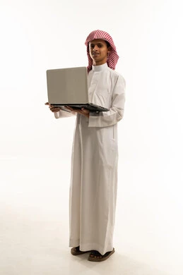 Looking at the camera with gestures of happiness and joy, the concept of distance education, a portrait of an Arab Gulf Saudi boy wearing traditional attire and a shemagh, using a laptop for research, reading, and browsing electronic applications, full-body, white background.