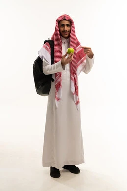 فاكهة طبيعية وطازجة, أهمية العناصر الغذائية الموجودة في التفاح, الطعام الصحي والمتوازن, بورتريه لطالب عربي خليجي سعودي يرتدي الزي التقليدي و الشماغ  و حقيبة الظهر يمسك تفاحة ، اتباع نظام غذائي، وجبة خفيفة لذيذة، الجسم بالطول الكامل، خلفية بيضاء