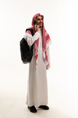 Making a voice call via mobile, enjoying exchanging conversations and news, a portrait of a Saudi Gulf Arab student wearing traditional attire and a shemagh with a backpack talking on a mobile phone, gestures of happiness while communicating with friends and relatives, full-body, white background.