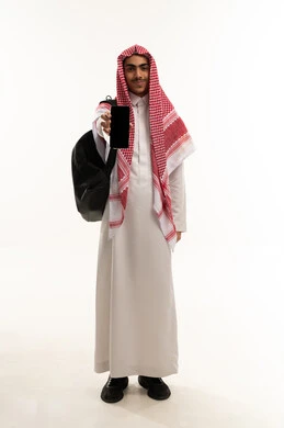 استخدام التقنيات والأجهزة الحديثة الالكترونية، إعلانات تسويقية ترويجية عبر الجوال، بورترية لطالب عربي خليجي سعودي يرتدي الثوب التقليدي و الشماغ و حقيبة الظهر يقوم بعرض الهاتف المحمول بشاشة سوداء فارغة ينظر الى الكاميرا بإيماءات السرور والسعادة، الجسم بالطول الكامل، خلفية بيضاء