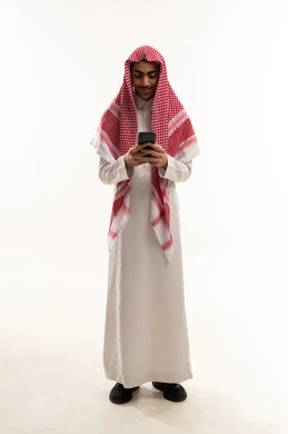 تصفح مواقع التواصل الاجتماعي، الاطمئنان على الاهل والاصدقاء , بورترية لصبي عربي خليجي سعودي يرتدي الثوب التقليدي و الشماغ يقوم باستخدام الهاتف المحمول, دمج التقنيات الحديثة فى الحياة اليومية, ايماءات السعادة و السرور، الجسم بالطول الكامل، خلفية بيضاء