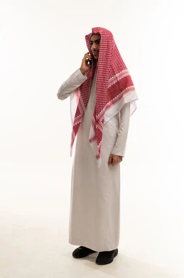 Enjoying exchanging conversations and news, making a voice phone call via mobile, a portrait of a Saudi Arabian Gulf boy wearing traditional attire and a ghutrah talking on a mobile phone, gestures of happiness while communicating with friends and relatives, full-body, white background.