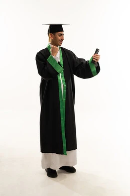 التفوق والنجاح والاجتهاد، توثيق لحظة النجاح عبر الهاتف المحمول، الاحتفال بإنهاء المرحلة الجامعية،  بورتريه لطالب عربي سعودي خليجي يرتدي العباءه وقبعة التخرج يقوم بالتقاط صور سيلفي بتعابير السرور، الجسم بالطول الكامل، خلفية بيضاء