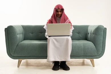 الاستمتاع بالمشاهدة عبر التقنية، الدراسة عن بعد من خلال الانترنت, إجراء مكالمة فيديو عبر جهاز الابتوب, بورتريه لفتى عربي خليجي سعودي يرتدي الثوب التقليدي يجلس على كنبه مريحة يقوم باستخدام الحاسب المحمول ، خلفية بيضاء