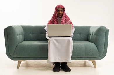إنجاز المهام المطلوبة عن بعد، دمج التكنولوجيا الحديثة في الحياة العملية, إجراء مكالمة فيديو عبر جهاز الابتوب, بورتريه لفتى عربي خليجي سعودي يرتدي الثوب التقليدي يجلس على كنبه مريحة يقوم باستخدام الحاسب المحمول ، خلفية بيضاء