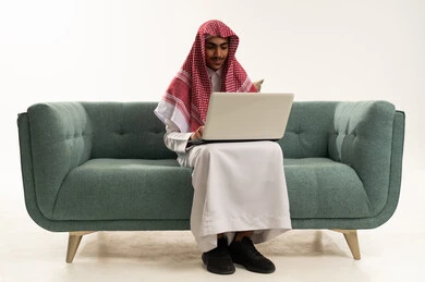 دمج التكنولوجيا الحديثة في الحياة العملية، الدراسة عن بعد من خلال الانترنت, إجراء مكالمة فيديو عبر جهاز الابتوب, بورتريه لفتى عربي خليجي سعودي يرتدي الثوب التقليدي يجلس على كنبه مريحة يقوم باستخدام الحاسب المحمول ، خلفية بيضاء