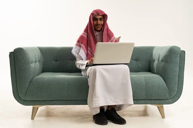 Distance learning through the internet, looking at the camera with gestures of happiness and joy, making a video call via a laptop, a portrait of an Arab Gulf Saudi boy wearing traditional attire sitting on a comfortable couch using a laptop, white background.