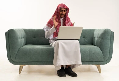 Raising the hand upwards with gestures of greeting and peace, making a video call via a laptop, gestures of joy and happiness, working remotely through the internet, a portrait of a Saudi Arabian Gulf boy wearing traditional attire sitting on a comfortable couch using a laptop, with a white background.