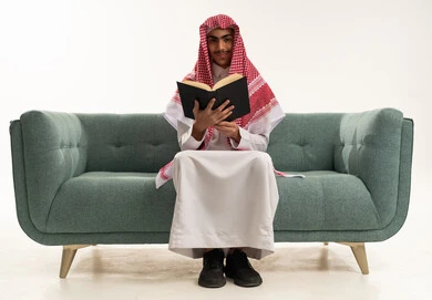 التركيز والاندماج بالمطالعة والقراءة, قضاء أوقات الفراغ بالتثقيف والتعلم, تنمية مهارات القراءة, الاهتمام بتصفح الكتب المفيدة, بورتريه لفتى عربي خليجي سعودي يرتدي الثوب التقليدي يجلس على كنبه مريحة ويمسك بيده الكتاب, خلفية بيضاء