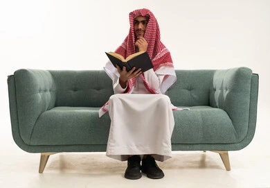 Finding a solution to a problem, the concept of daydreaming, contemplation, and planning for the future, a portrait of a young Arab Gulf Saudi man wearing traditional attire and a ghutrah sitting on a sofa reading a book and resting his hand on his chin with gestures of thought and confusion, white background.