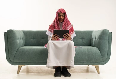 ايماءات الاندماج و التركيز، التواصل و الدردشة مع الاقارب, القيام بمقابلات أونلاين، استخدام جهاز تقني حديث لتصفح الصور والفيديوهات بسهولة، بورتريه لشاب عربي خليجي سعودي يرتدي الثوب الاتقليدي و الشماغ يجلس على الاريكة يمسك بيده جهاز التابلت , خلفية بيضاء