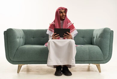 Looking at something, communicating and chatting with relatives, conducting online interviews, using a modern technical device to easily browse photos and videos, a portrait of a young Arab Gulf Saudi man wearing traditional attire and a ghutrah sitting on a sofa holding a tablet in his hand, white background.