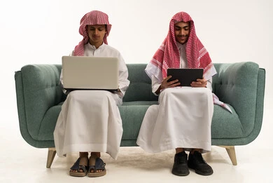 Spending enjoyable times on the tablet, the concept of online learning, conversations and chatting over the internet, communicating with friends from a distance, a portrait of an Arab Saudi Gulf boy wearing the Saudi thobe and shemagh sitting on the couch with a friend using a laptop, white background.