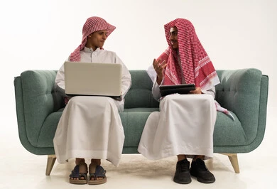 محادثات ودردشة عبر الانترنت، التواصل مع الأصدقاء عن بعد, بورتريه لولد عربي سعودي خليجي يرتدي الثوب السعودي و الشماغ يجلس على الاريكة برفقة صديقة يقوم بإستخدام جهاز الحاسوب المحمول، مفهوم التعلم اونلاين، قضاء أوقات ممتعة على التابلت، خلفية بيضاء