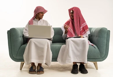 التواصل مع الأصدقاء عن بعد، محادثات ودردشة عبر الانترنت, بورتريه لولد عربي سعودي خليجي يرتدي الثوب السعودي و الشماغ يجلس على الاريكة برفقة صديقة يقوم بإستخدام جهاز الحاسوب المحمول، مفهوم التعلم اونلاين، قضاء أوقات ممتعة على التابلت، خلفية بيضاء