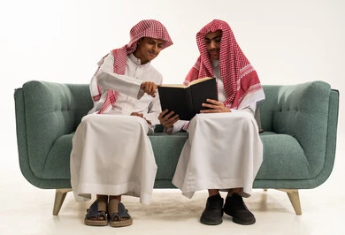 الاستمتاع بقراءة الكتب ، قضاء الوقت بالقراءة والتعلم والتثقف، بورتريه لولدان عربيان سعوديان خليجيان يرتديان الزي السعودي و الشماغ  يجلسان على الاريكة يقومان بمشاركه قراءة كتاب , خلفية بيضاء