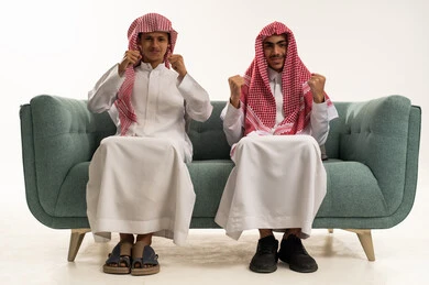 النظر الى الكاميرا بتعابير السرور و السعادة، ايماءات الفرح والانتصار، بورتريه لولدان عربيان خليجيان سعوديان يرتديان الزي التقليدي و الشماغ يجلسان على الاريكة يرفعان  قبضة يدها للاعلى بحركات الفوز، خلفية بيضاء