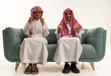 ايماءات الفرح والانتصار، بورتريه لولدان عربيان خليجيان سعوديان يرتديان الزي التقليدي و الشماغ يجلسان على الاريكة يرفعان  قبضة يدها للاعلى بحركات الفوز، النظر الى الكاميرا بتعابير السرور و السعادة، خلفية بيضاء