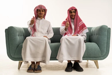 تعابير الإعجاب والرضا، مفهوم القبول والموافقه، بورتريه لولدان عربيان خليجيان سعوديان يرتديان الزي التقليدي و الشماغ يجلسان على الاريكة يرفعان اصبع الابهام بإيماءات الامتياز والجودة، خلفية بيضاء