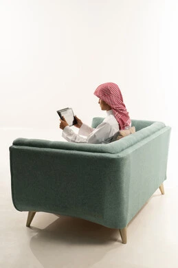 متابعة منصات التعليم الالكتروني المتاحة عبر الانترنت، مشاهده الافلام و المسلسلات عبر الجهاز اللوحي ، عرض بث مباشر على إحدى وسائل السوشل ميديا، إجراء مكالمة فيديو عبر التابلت، بورتريه من الجانب لشاب عربي خليجي سعودي يرتدي الثوب الابيض و الشماغ يجلس على الاريكة يقوم بالتواصل اونلاين مع الاقارب والاصدقاء، خلفية بيضاء