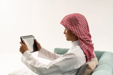 Watching movies and series on a tablet, live streaming on one of the social media platforms, making a video call via the tablet, a side portrait of a young Arab Gulf Saudi man wearing a white thobe and a shemagh sitting on the sofa communicating online with relatives and friends, following available online educational platforms, white background.