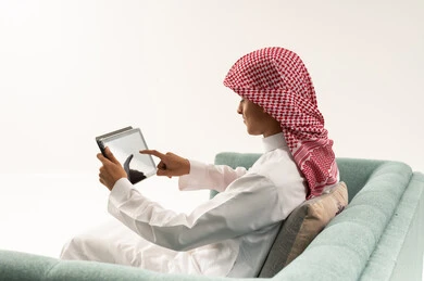Tapping on the tablet screen, watching a live broadcast on one of the social media platforms, making a video call via the tablet, a side portrait of a young Arab Gulf Saudi man wearing a white thobe and a shemagh sitting on the couch communicating online with relatives and friends, following available online educational platforms, white background.