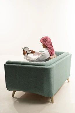 A live broadcast on one of the social media platforms, making a video call via a tablet, a side portrait of a young Arab Gulf Saudi man wearing a white thobe and a shemagh sitting on the couch communicating online with relatives and friends, following available online educational platforms, white background.