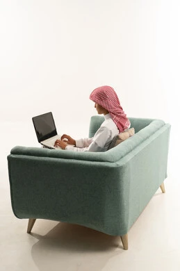 النقر على لوحة المفاتيح، التعلم عن بعد وتلقي الدروس عبر الأونلاين، أنشطة ترفيهية الكترونية مسلية، استخدام التكنولوجيا والتقنية الحديثة والمتطورة، بورتريه من الجانب لشاب عربي خليجي سعودي يرتدي الثوب الابيض و الشماغ يجلس على الاريكة يستخدم جهاز الحاسوب المحمول ، خلفية بيضاء