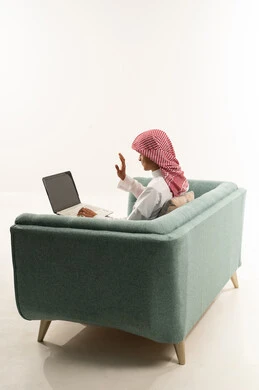 إجراء مكالمة فيديو باستخدام الحاسب المحمول، رفع كف اليد دليل على القاء التحية ورد السلام، تبادل الأخبار مع الأهل والأصدقاء، بورتريه من الجانب لشاب عربي خليجي سعودي يرتدي الثوب الابيض و الشماغ يجلس على الاريكة يقوم بالتواصل مع الأقارب، تعابير الفرح والسرور، خلفية بيضاء